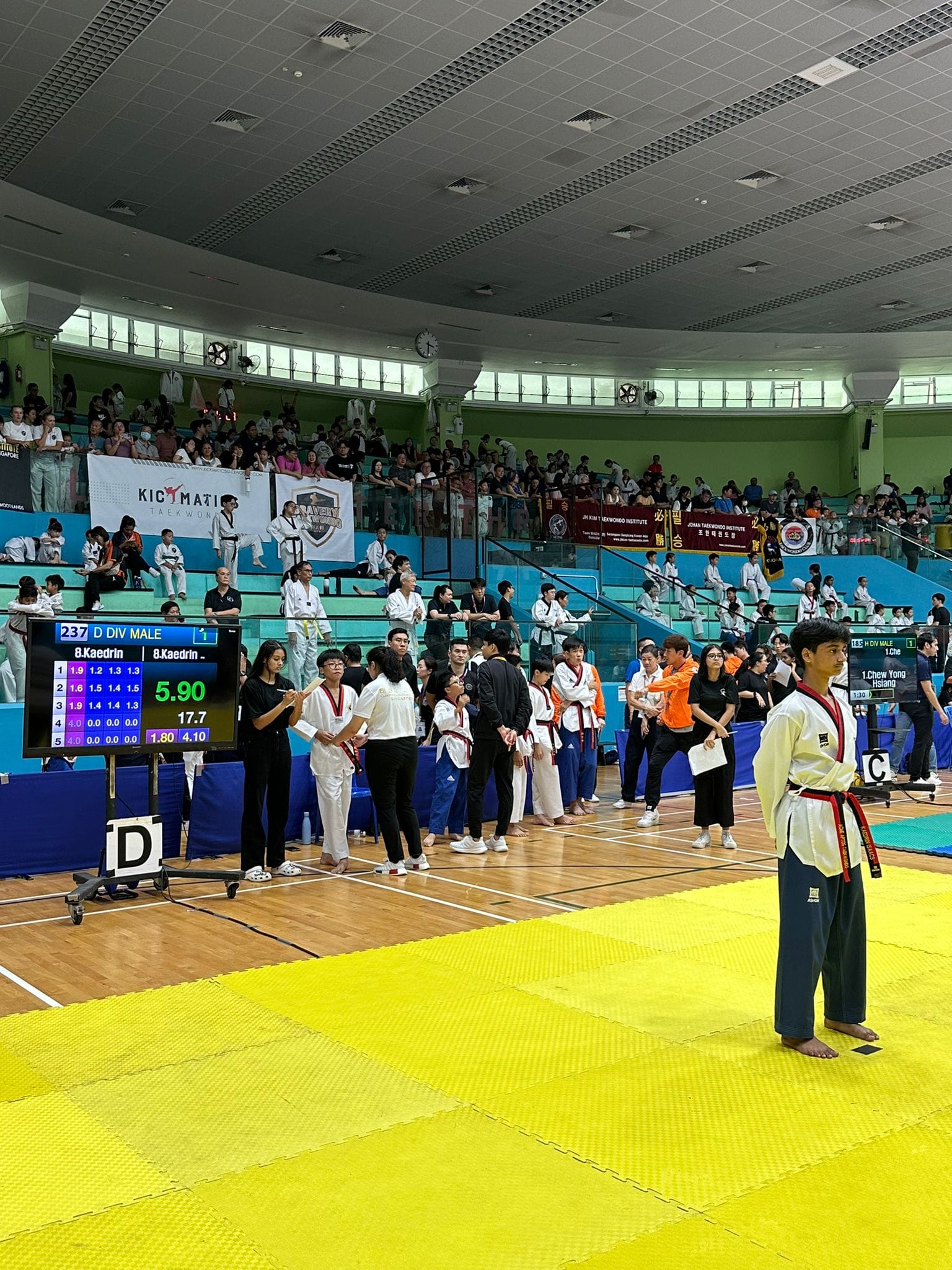 A taekwondo competitor stands on yellow mats as a scoreboard displays 'D DIV MALE, 8.Kaedrin, 5.90' in a crowded competition hall.